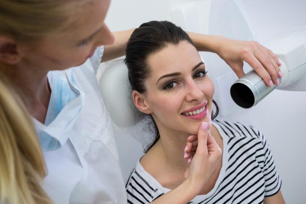 Dentist gently examining a smiling female patient’s jaw while preparing dental equipment in a clinical setting.